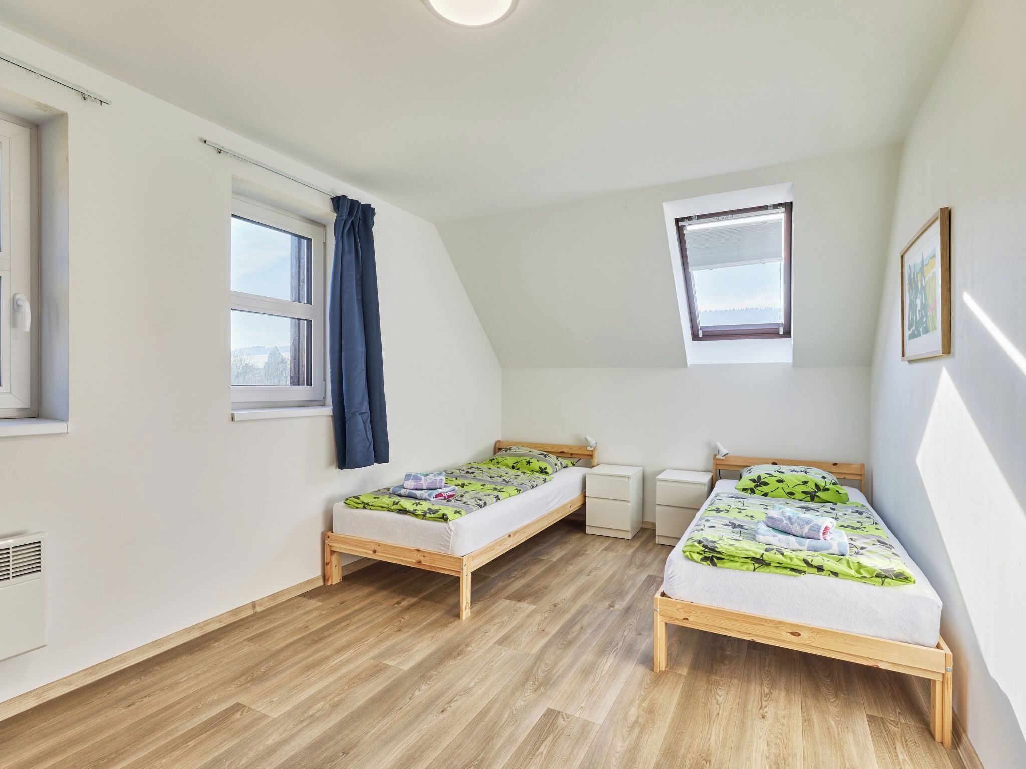 Une chambre à coucher avec deux lits en bois, une fenêtre avec des rideaux, un ventilateur de plafond et un tableau encadré accroché au mur.