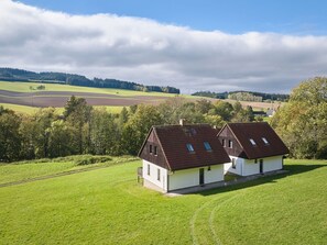 Kaksi modernia taloa, joissa on kaltevat katot ja kattovalokuviot, ja niiden ympärillä on vehreitä niittyjä ja taustalla metsäinen kukkula.