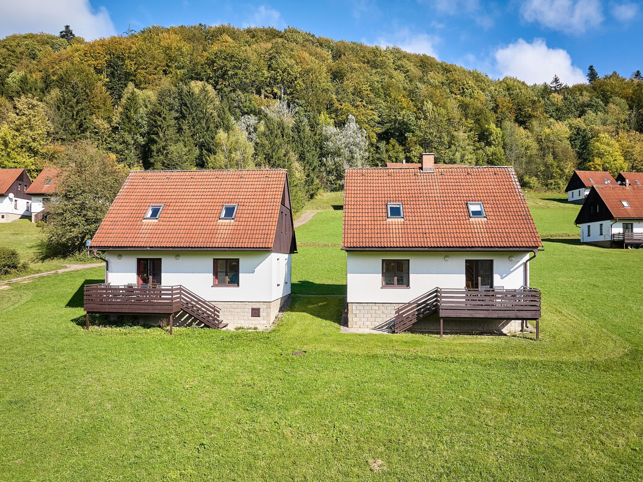 Twee witte huizen met rode daken en houten balkons, omringd door groen gras en bomen.