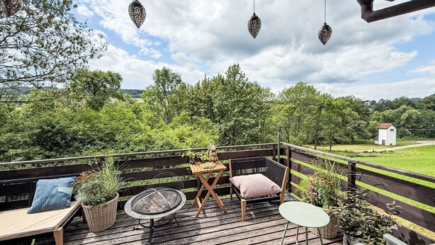 A wooden deck with outdoor furniture, including a table and chairs, surrounded by greenery and a view of the countryside.