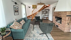 A spacious living room with a teal sofa, a grey armchair, a wooden staircase, and a brick fireplace.