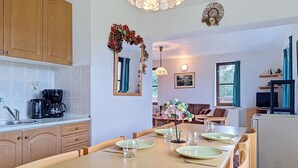 Et moderne køkken med træskabe, et spisebord dækket med tallerkener og glas, samt en opholdsstue med sofa og fjernsyn.