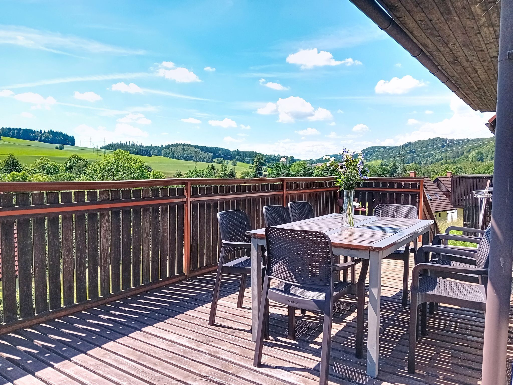 Eine Holzterrasse mit Tisch und Stühlen, die auf eine grüne Landschaft mit Hügeln blickt.
