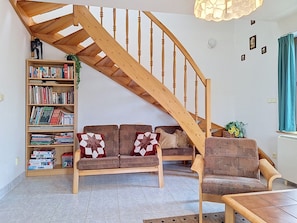 A living room with a wooden staircase, a sofa with patterned cushions, a bookshelf, and a small table.