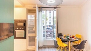 A compact kitchen with modern appliances, a dining area with yellow chairs, and a view of the city through a balcony.