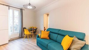 A living room with a teal sofa, yellow cushions, a dining table with chairs, and a large window.