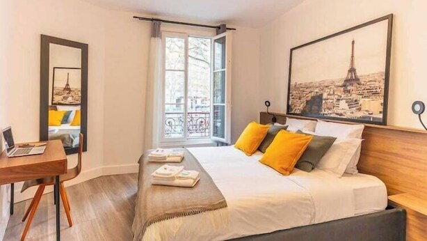 A hotel room with a bed, a desk, a chair, a laptop, a window with a view of buildings, and a framed picture of a cityscape.