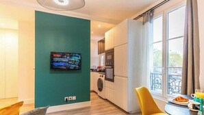 A modern kitchen with a washing machine, a dining area with yellow chairs, and a TV mounted on a teal wall.