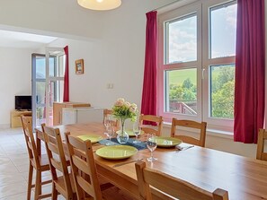 A dining area with a wooden table set for four, surrounded by chairs, with a view of greenery outside through large windows.