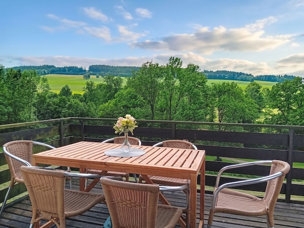 A wooden deck with a table and wicker chairs, overlooking a green landscape.