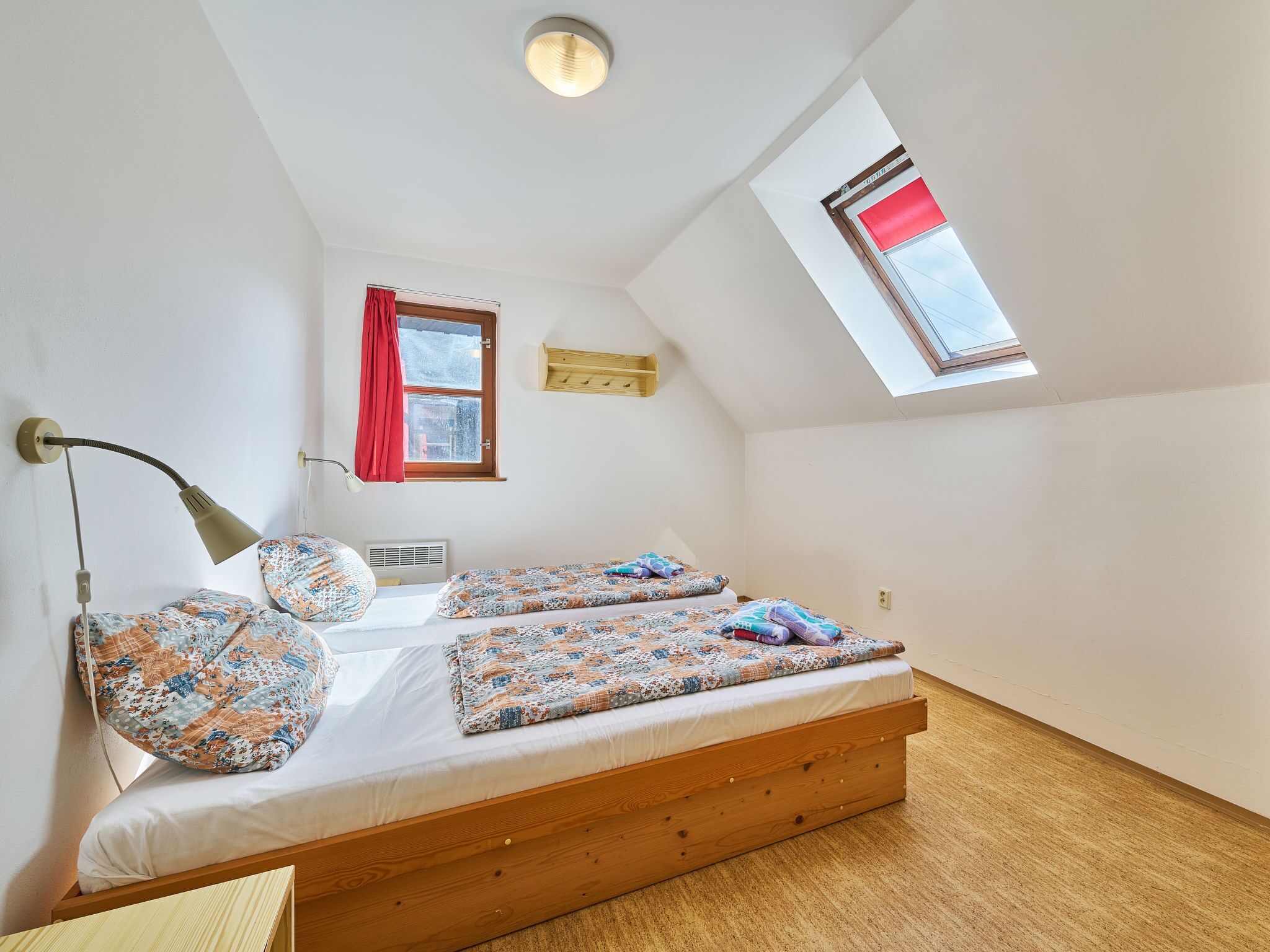 Een slaapkamer met een houten bed, twee kussens, een bedlampje, een raam met rood gordijn en een dakraam.