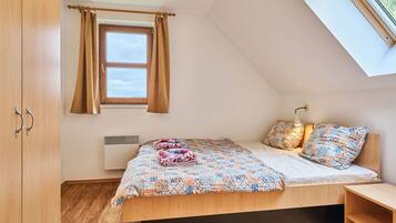 Un dormitorio con techo inclinado, una cama con cubrecama floral, dos almohadas, una mesita de noche y una ventana con cortinas.