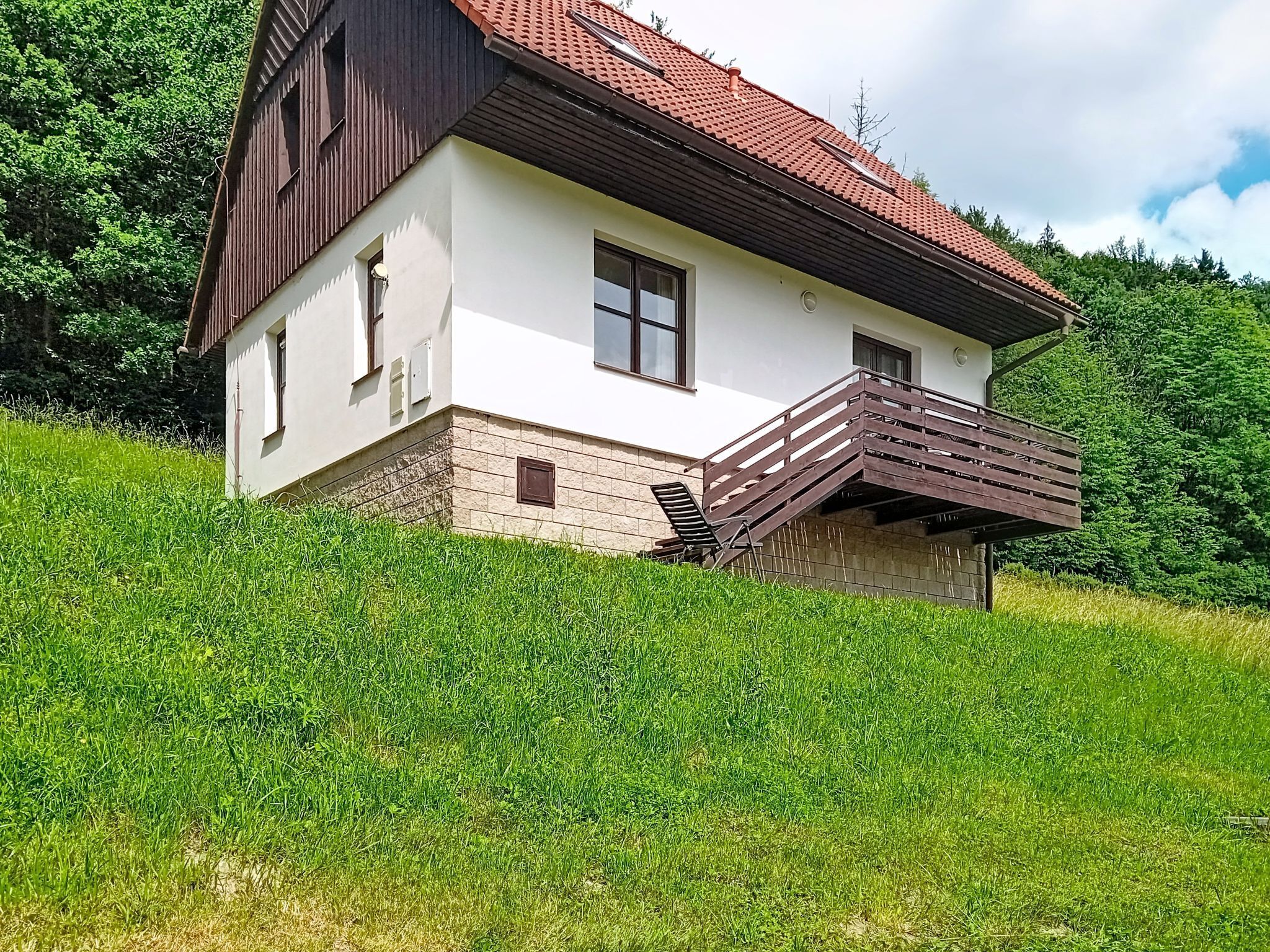A two-story house with a balcony, situated on a grassy hill with trees in the background.