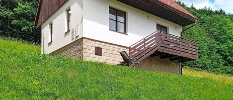 A two-story house with a balcony, situated on a grassy hill with trees in the background.