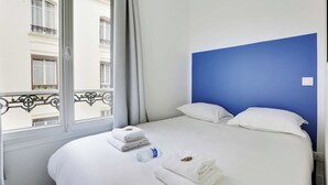 A hotel room with a bed, white towels, a bottle of water, and a view of a building through a window.