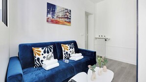 A modern living room with a blue sofa, white cushions, a painting of a cityscape, a radiator, and a small table with decorative items.