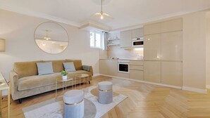 Un salon moderne avec un canapé, une table basse et des poufs, une cuisine équipée de meubles et d’appareils électroménagers, et un grand miroir accroché au mur.