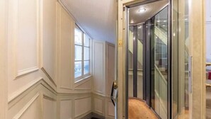 A hallway with a glass elevator door, a carpeted staircase, and a window with a view of the outside.