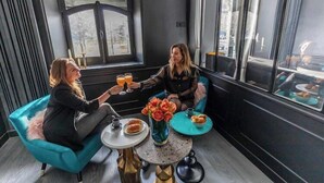 Two people sitting on a teal sofa, toasting with drinks, with a window view and a small table with a vase of flowers and a plate of food.