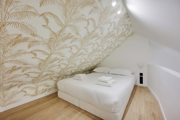 Un dormitorio con una cama, ropa de cama blanca, una pared con diseño de hojas y suelo de madera.