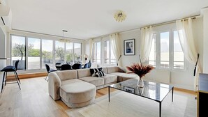A modern living room with a sectional sofa, a glass coffee table, and a dining area with a table and chairs. Large windows provide natural light and views of the outdoors. - Stunning apartment - 3BR/6P - La DÃ©fense (Courbevoie)