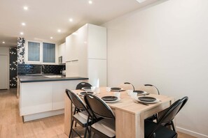 A modern kitchen with white cabinets, a black countertop, and a dining area with a wooden table and black chairs. - Spacious apartment with 2 rooms (Paris)