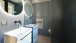 A modern bathroom with a white sink, a blue cabinet, a large mirror with a circular light, and a shower with a handheld showerhead.