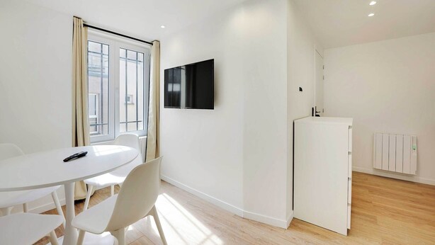 A modern, minimalistic room with a white table, chairs, a flat-screen TV mounted on the wall, and a white cabinet.