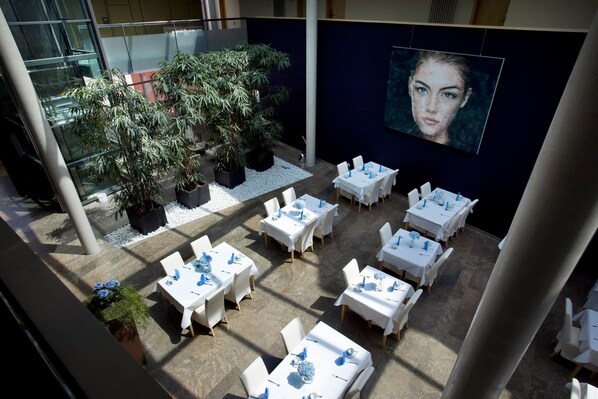 An indoor dining area with tables set for guests, surrounded by potted plants and a large wall mural.