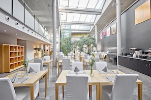A modern restaurant with a high ceiling and skylights, featuring a central dining area with tables and chairs, a kitchen area on the right, and a bookshelf on the left.