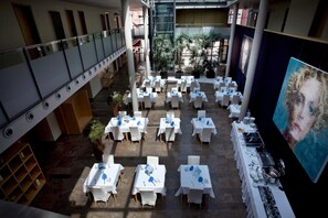 A spacious indoor area with multiple tables set for dining, a large painting on the right, and a balcony with plants.