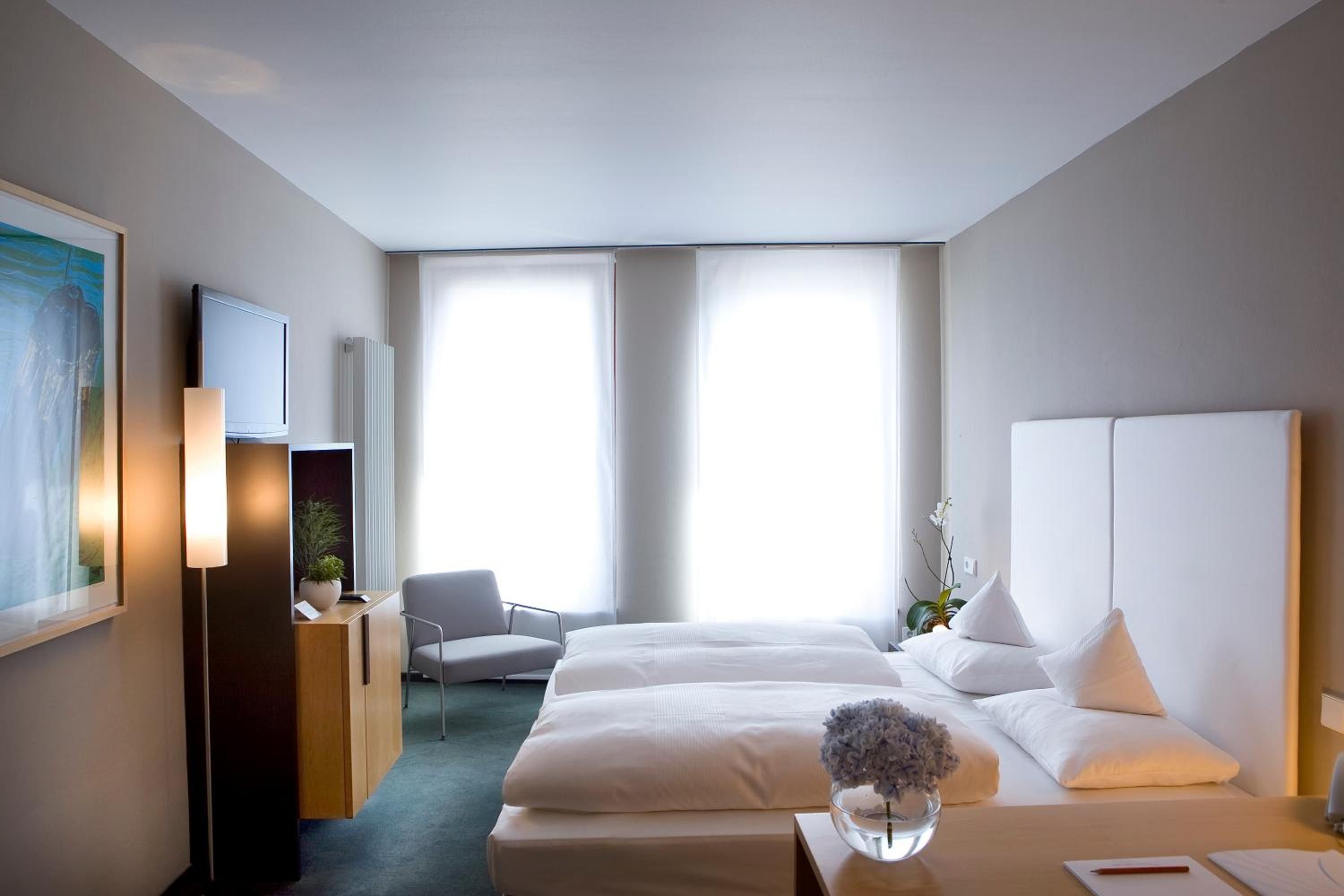 A hotel room with a bed, bedside table, lamp, chair, TV, and a large window.