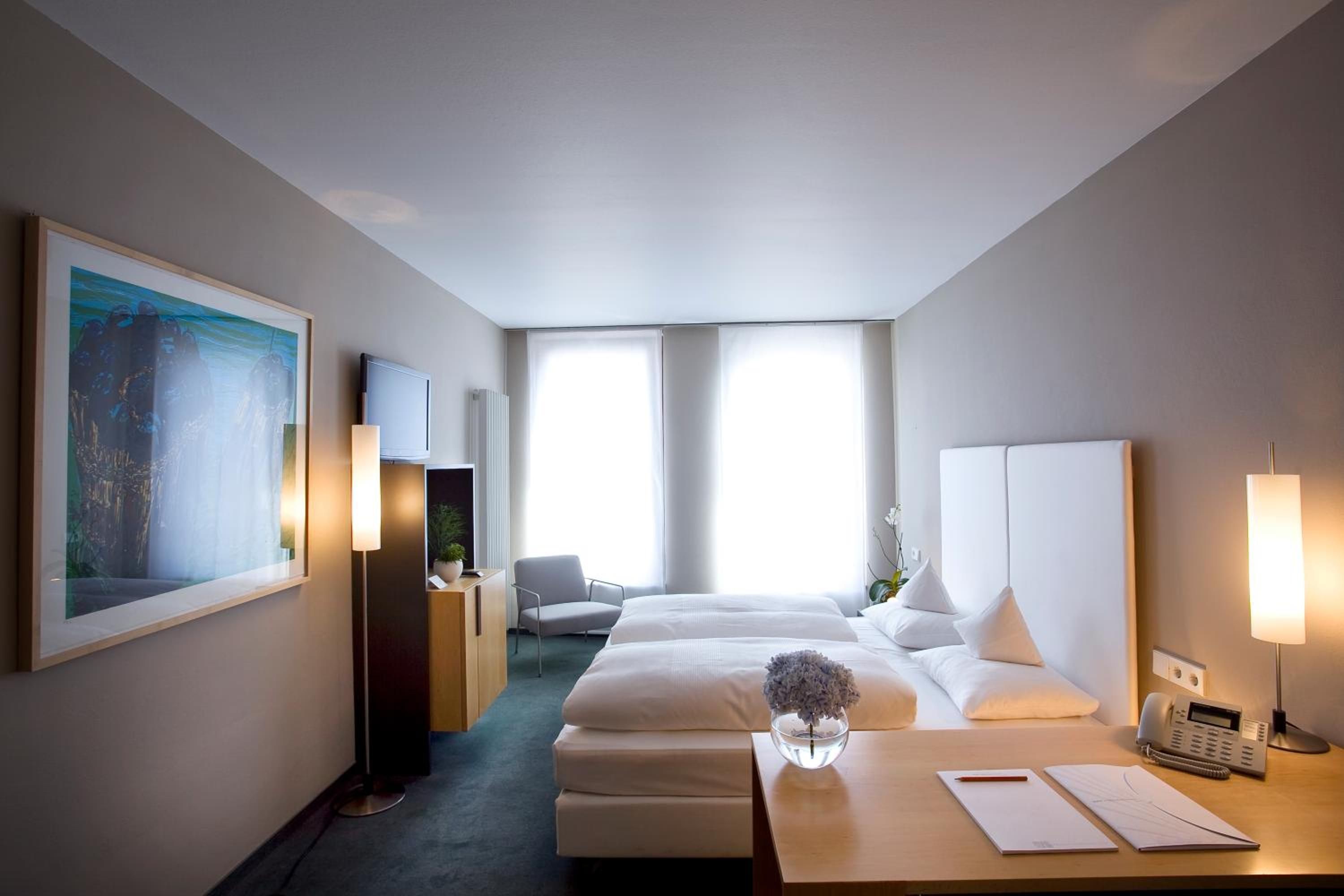 A hotel room with two beds, a desk with a telephone and papers, a chair, a large painting, and a radiator.
