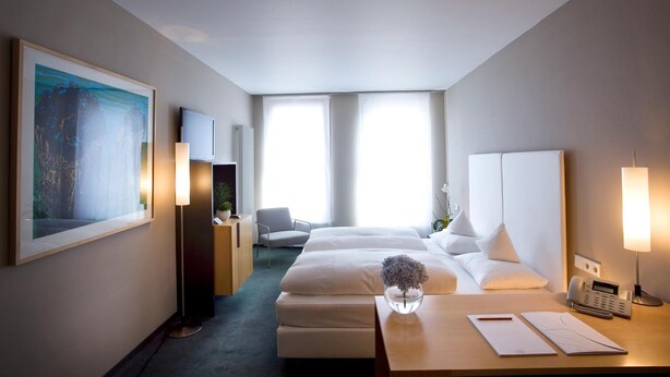 A hotel room with two beds, a desk with a telephone and papers, a chair, a large painting, and a radiator.