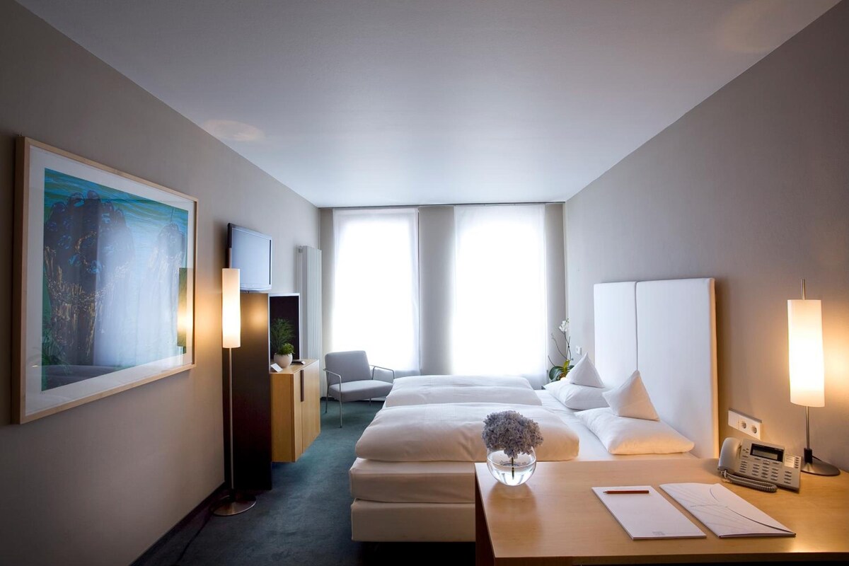 Habitació d'hotel amb dos llits, una taula de treball amb un telèfon i papers, una cadira, un quadre gran i un radiador.