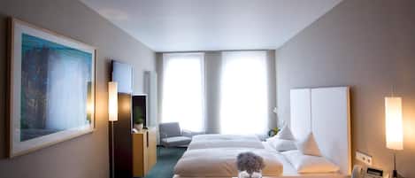 A hotel room with two beds, a desk with a telephone and papers, a chair, a large painting, and a radiator.