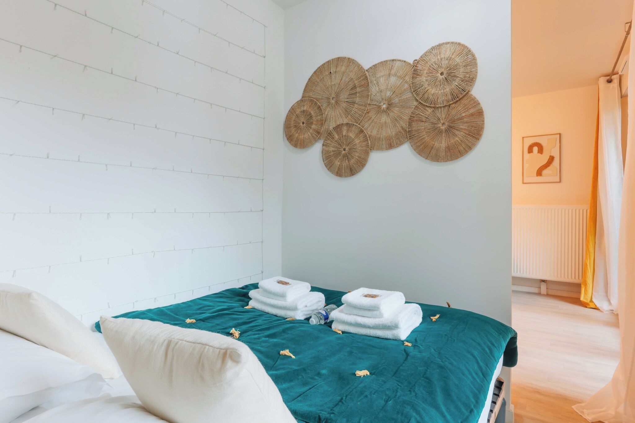 A hotel room with a bed, teal bedspread, white pillows, and a wall with woven wall art.