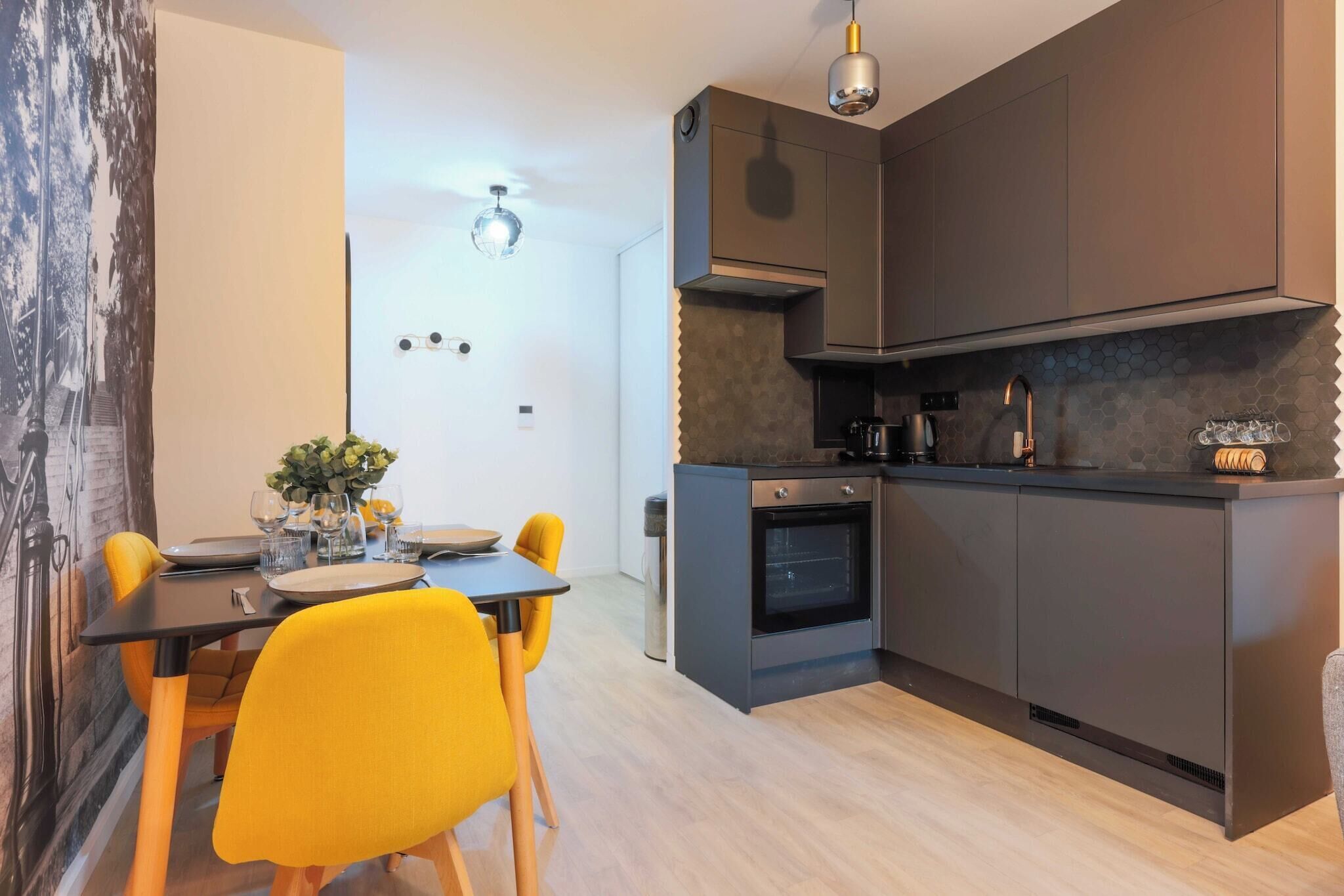 A modern kitchen with dark cabinetry, a built-in oven, and a dining area with a table and yellow chairs.