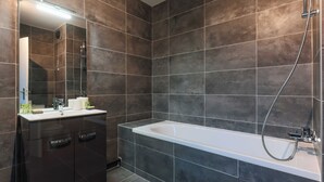 Uma casa de banho moderna com azulejos escuros, uma banheira branca e um lavatório com espelho.