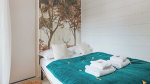 Cama bem arrumada com colcha azul-esverdeada e almofadas brancas, um grande mural de árvores na parede e uma parede de tijolos brancos.