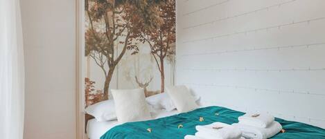 A neatly made bed with a teal bedspread and white pillows, a large wall mural of trees, and a white brick wall.