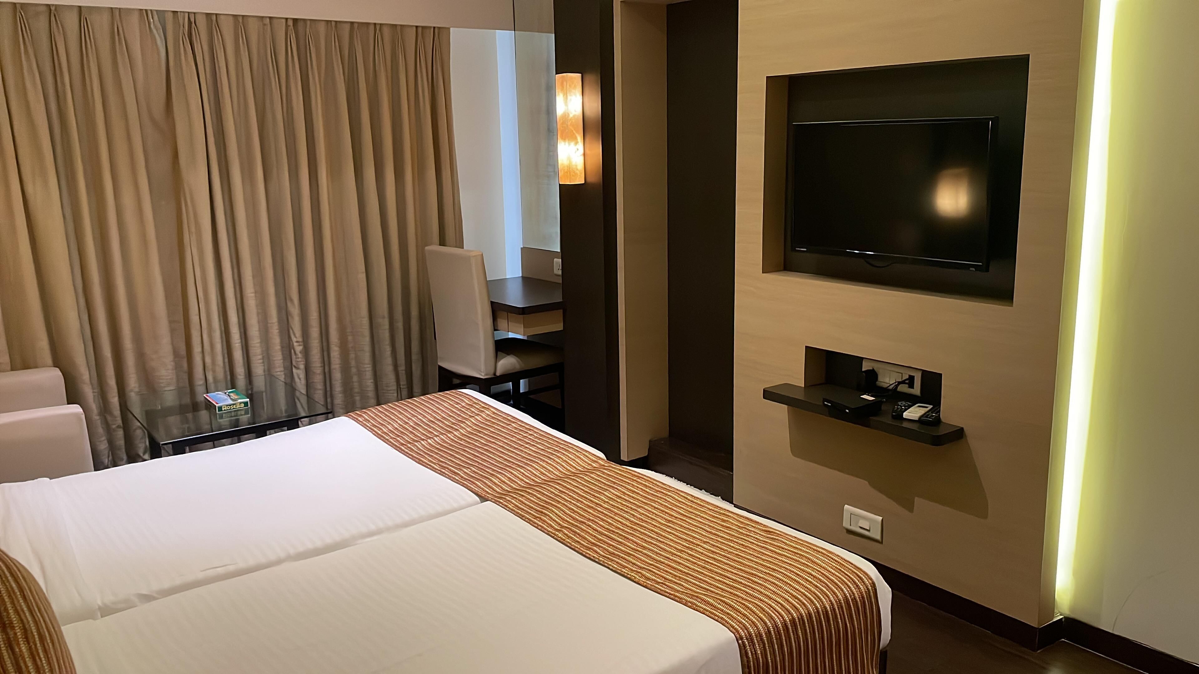 Habitació d'hotel amb un llit, televisió penjada a la paret, escriptori i cadira, i una cortina.