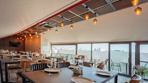 Un restaurant spacieux, doté de nombreuses tables et chaises, de grandes fenêtres donnant sur un plan d’eau, et d’un plafond moderne au design original, avec des poutres apparentes et des luminaires suspendus.