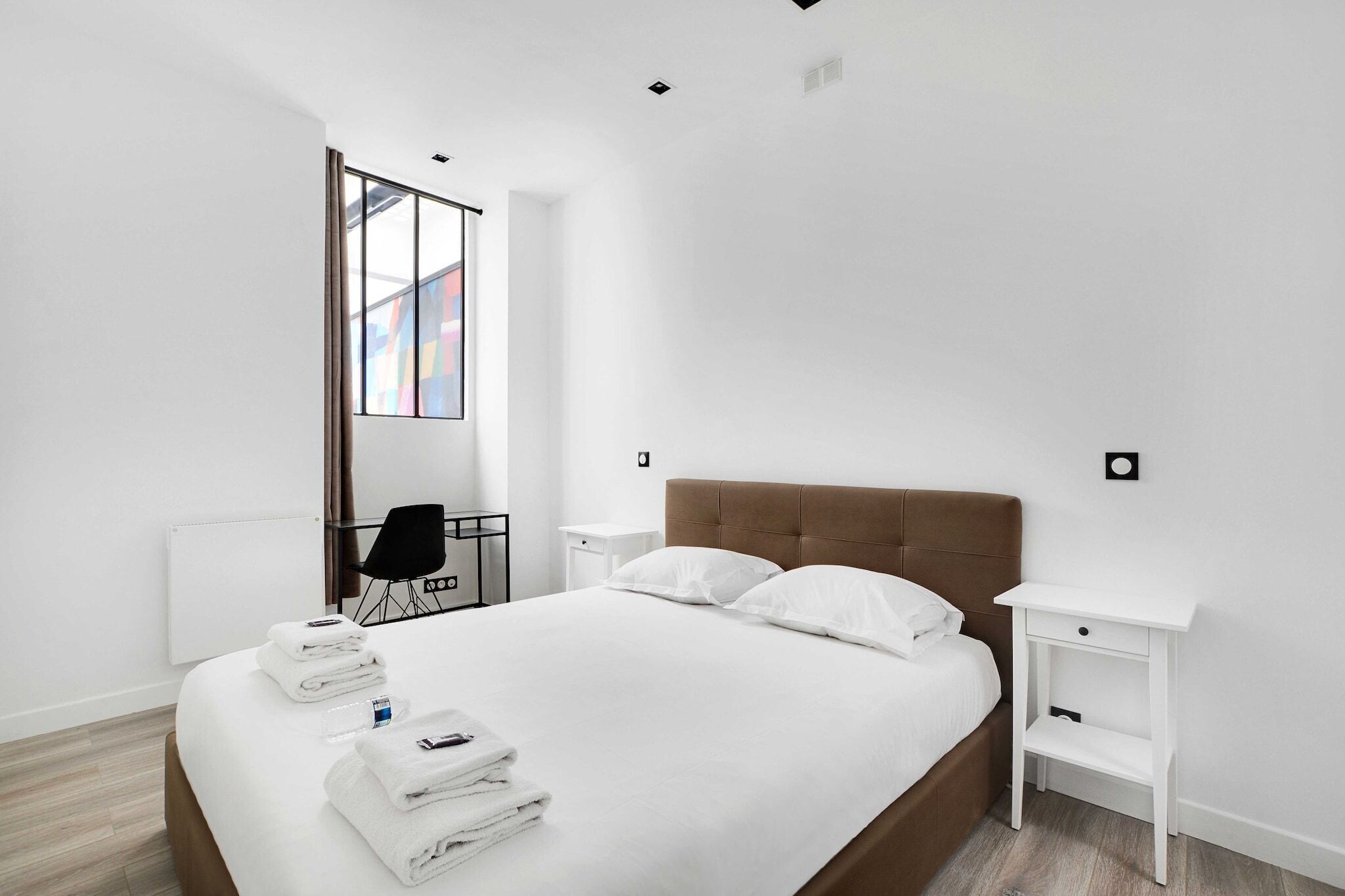 Habitación de hotel moderna con una cama grande, ropa de cama blanca, una mesita de noche y vistas a un edificio colorido.