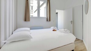 Un lit bien fait, avec du linge de lit blanc, une fenêtre avec des rideaux, une porte et un miroir rond accroché au mur.