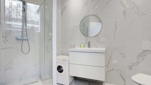 Une salle de bain moderne dotée d’une douche à parois de verre, d’un lave-linge encastré et d’un meuble-lavabo blanc avec un miroir rond.