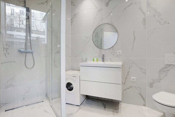 A modern bathroom with a glass-enclosed shower, a built-in washing machine, and a white vanity with a round mirror. - Intimate apartment - 1BR/4P - Saint-Lambert (Paris)