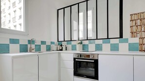 Une cuisine avec des meubles blancs, un four noir et une fenêtre donnant sur des immeubles.