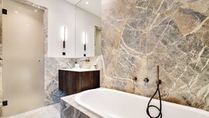 A modern bathroom with a marble wall, a freestanding bathtub, a vanity with a dark wood cabinet, and a large mirror.
