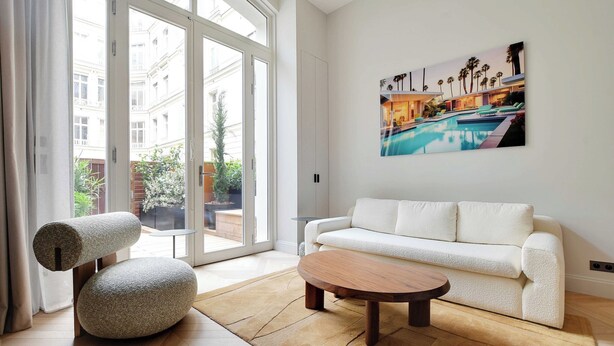 A modern living room with a white sofa, a round wooden coffee table, and a stone pouf. A large window offers a view of a pool and palm trees.